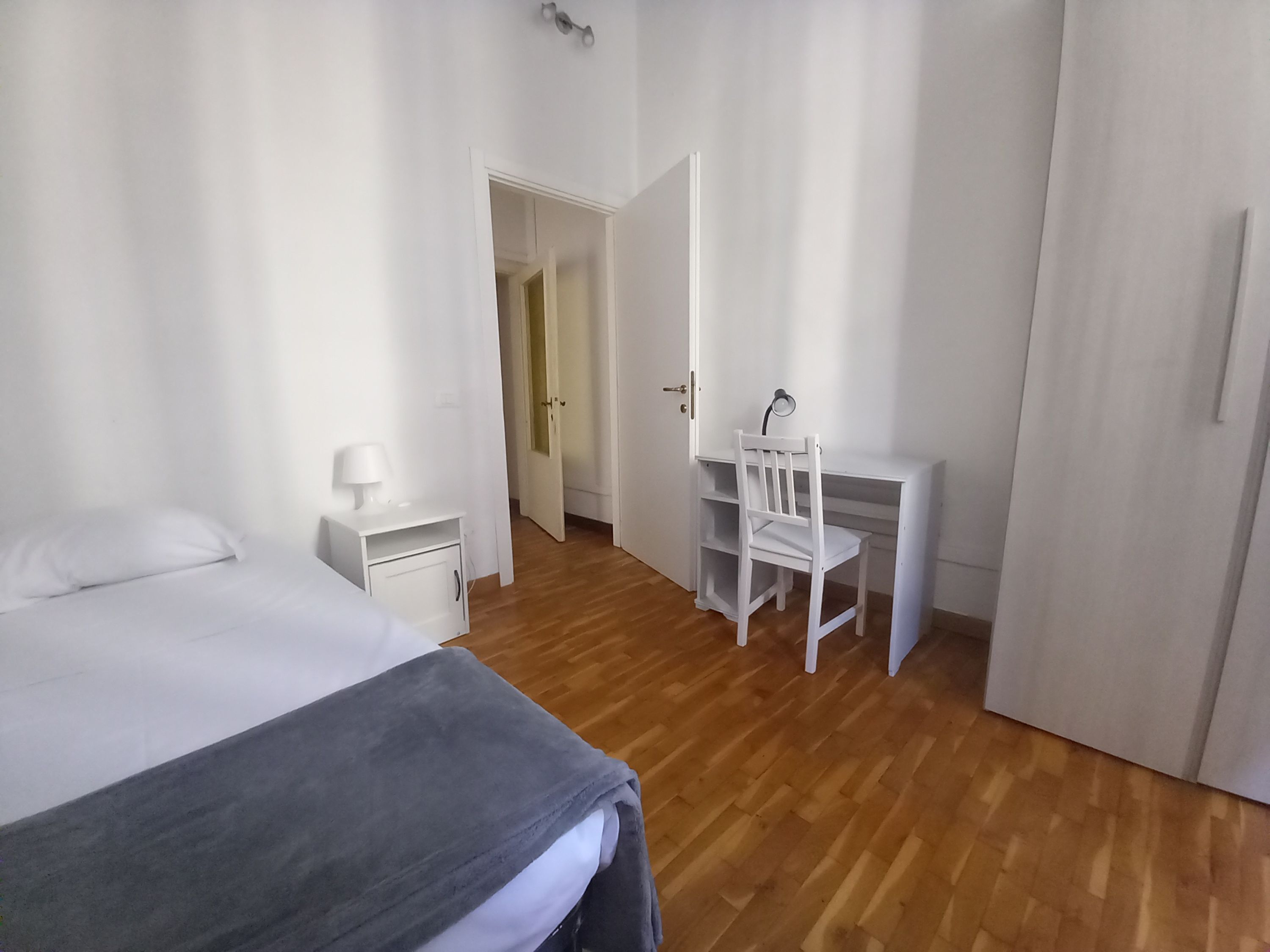 A bedroom with a bed, a desk, and a chair. There is a nightstand with a lamp, and a door leading to another room.