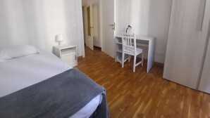 Quarto com cama, escrivaninha e cadeira. Tem uma mesa de cabeceira com abajur, e uma porta que dá para outro cômodo.