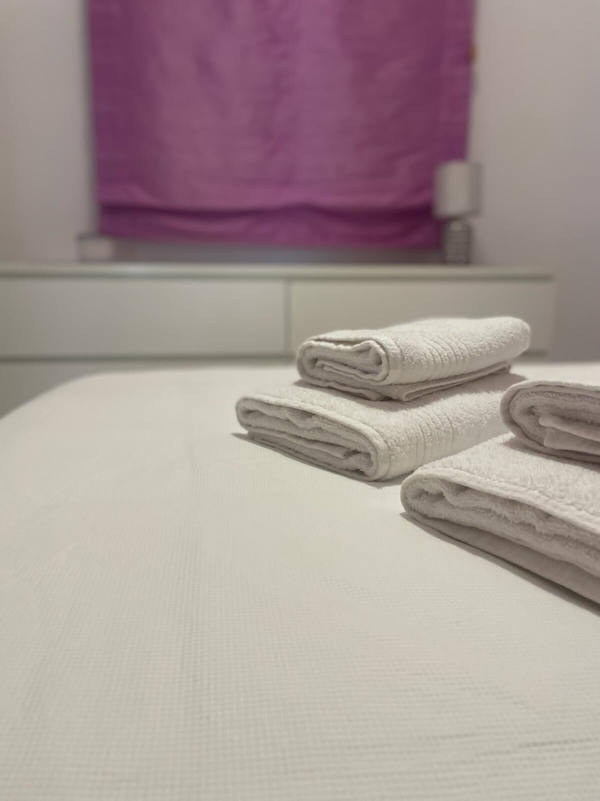 A neatly made bed with folded white towels and a purple towel hanging in the background.