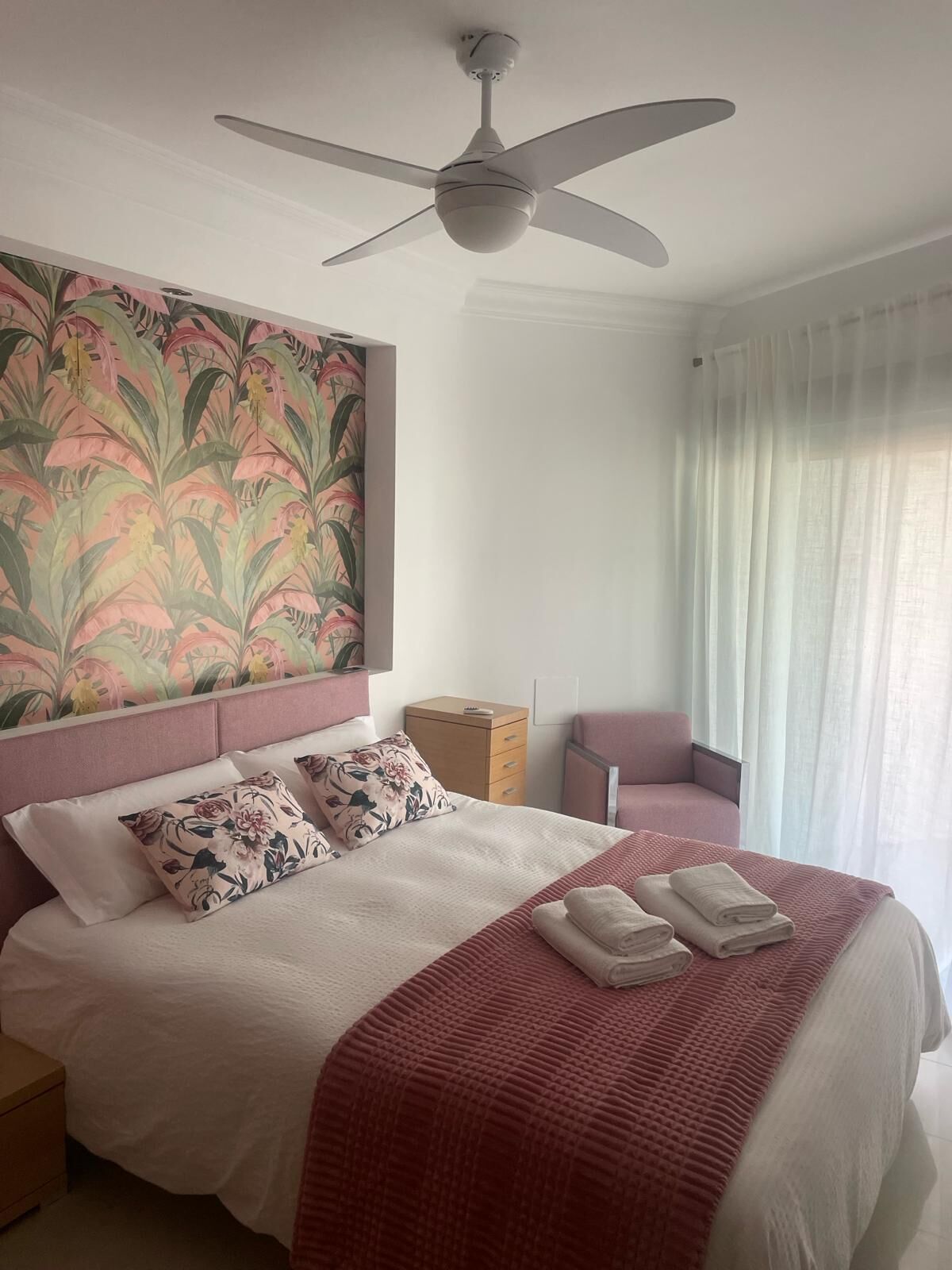 Ein ordentlich gemachtes Bett mit roter Decke und Kissen, ein Deckenventilator, ein Nachttisch, ein Holzkabinet und ein Fenster mit durchsichtigen Vorhängen.