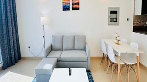 A modern living room with a grey sofa, a white coffee table, and a dining area with a white table and chairs. There are two wall-mounted light fixtures and two framed pictures on the wall. - OYO 1194 Home Orion Building 514 (Dubai)