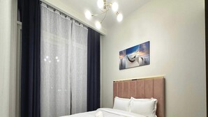 Un lit bien fait, avec du linge de lit blanc et une serviette enroulée, une grande fenêtre avec des rideaux foncés, un luminaire au plafond et une œuvre d’art accrochée au mur représentant un bateau.