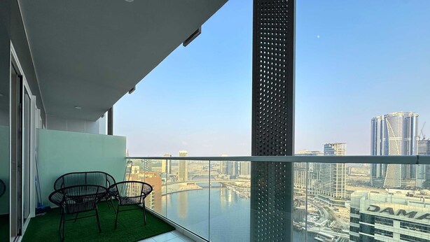 Un balcon doté d’une balustrade en verre, offrant une vue sur le paysage urbain, notamment sur une rivière et des immeubles de grande hauteur.
