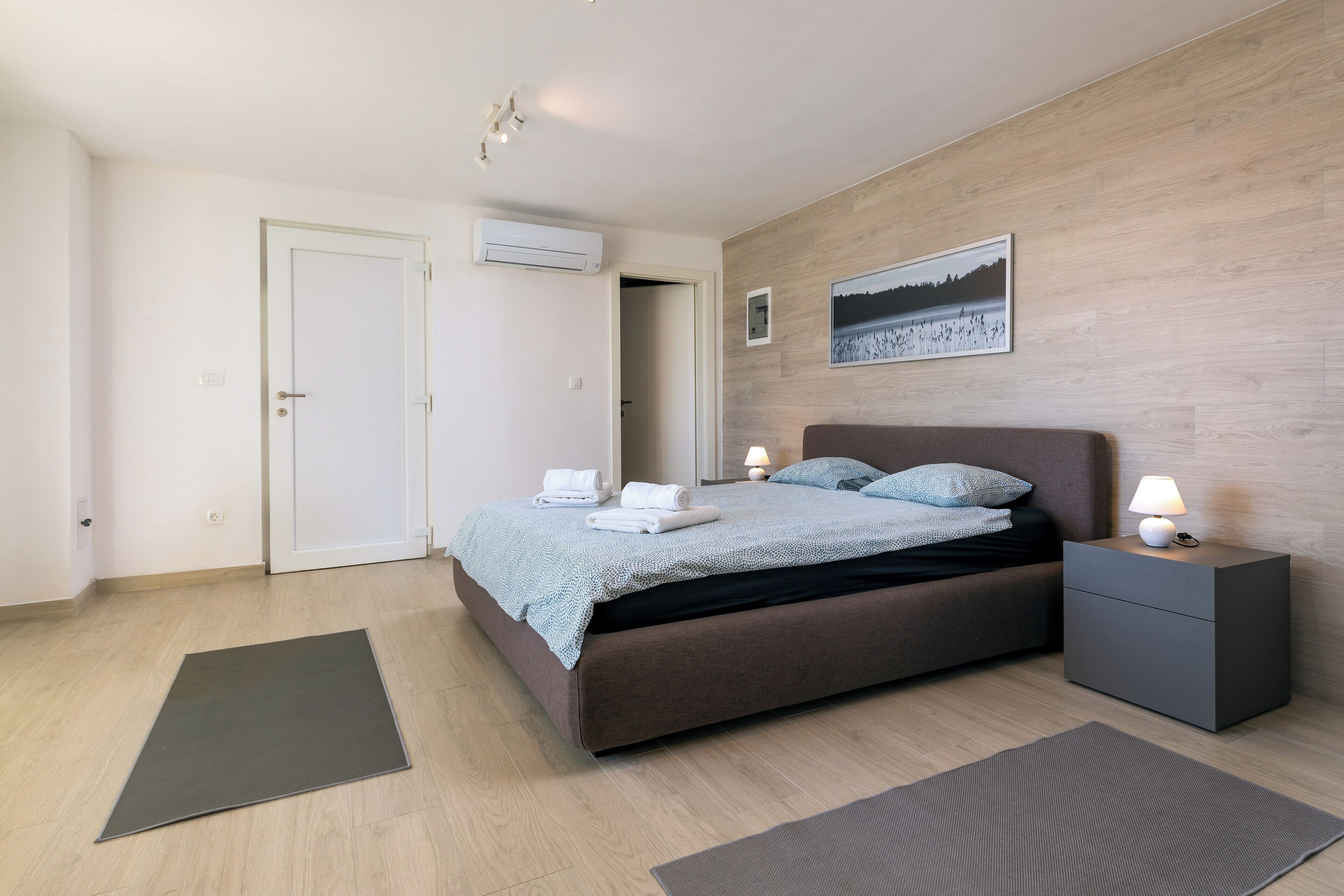A modern bedroom with a bed, bedside tables, a nightstand, a wall-mounted air conditioner, and a framed picture.