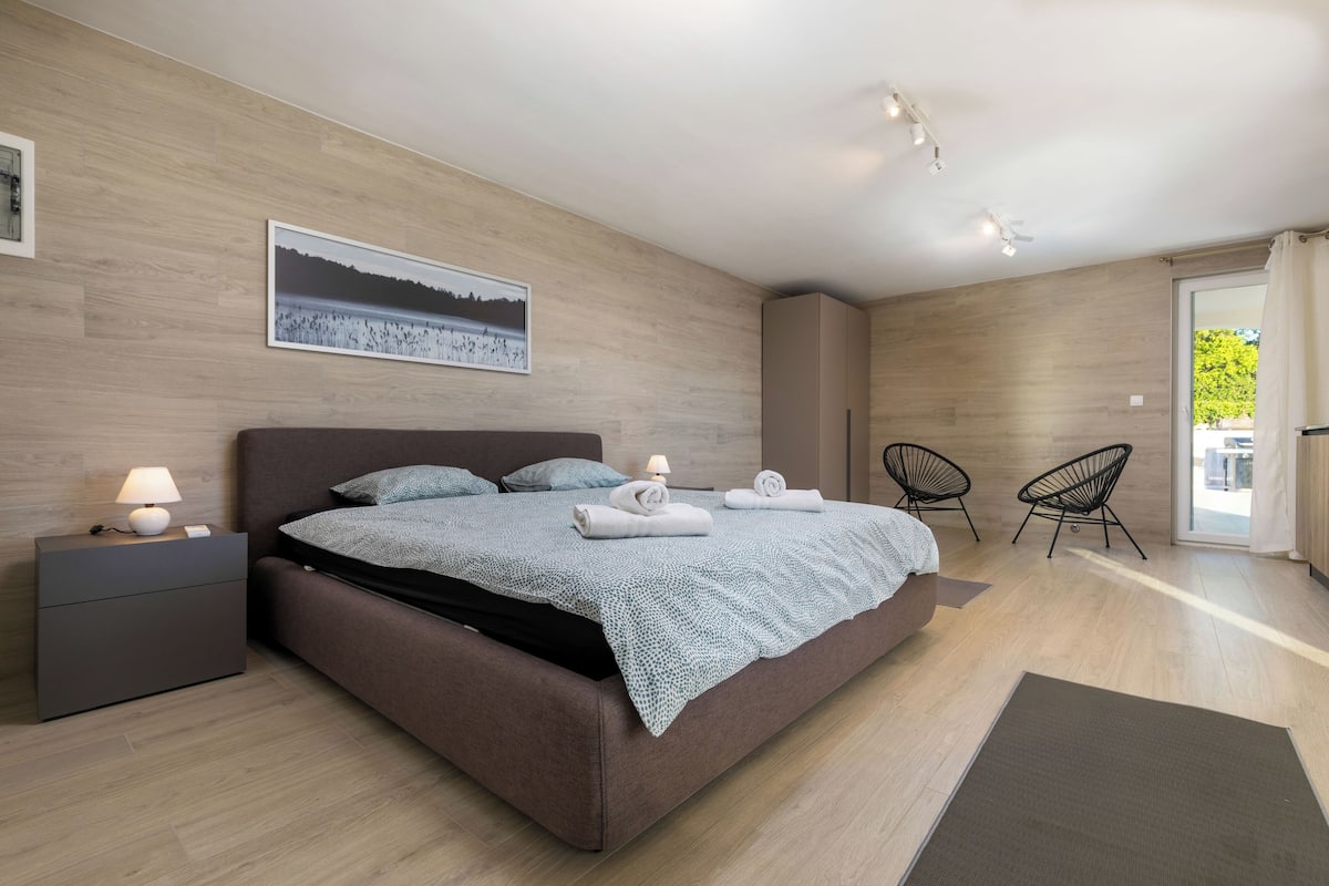 A modern bedroom with a large bed, a nightstand with a lamp, a chair, and a view of the outdoors.