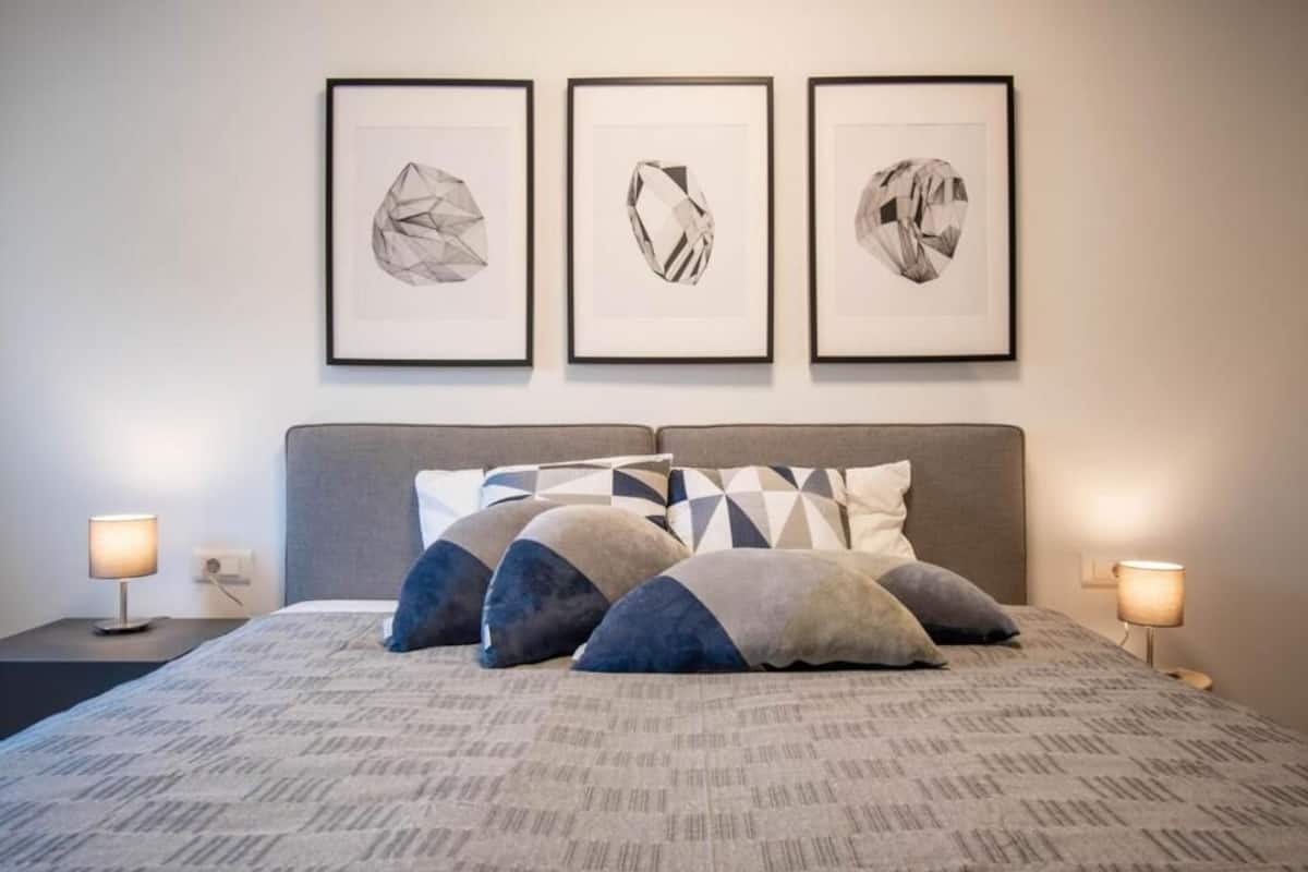 A neatly made bed with a patterned bedspread, a grey headboard, and three framed abstract artworks above it.