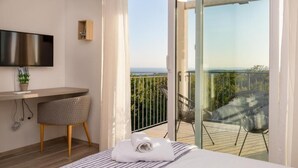 Quarto de hotel moderno com cama, escrivaninha, cadeira, TV e varanda com vista para o mar.