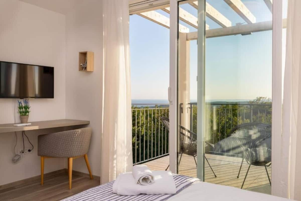 A modern hotel room with a bed, a desk, a chair, a TV, and a balcony with a view of the sea.