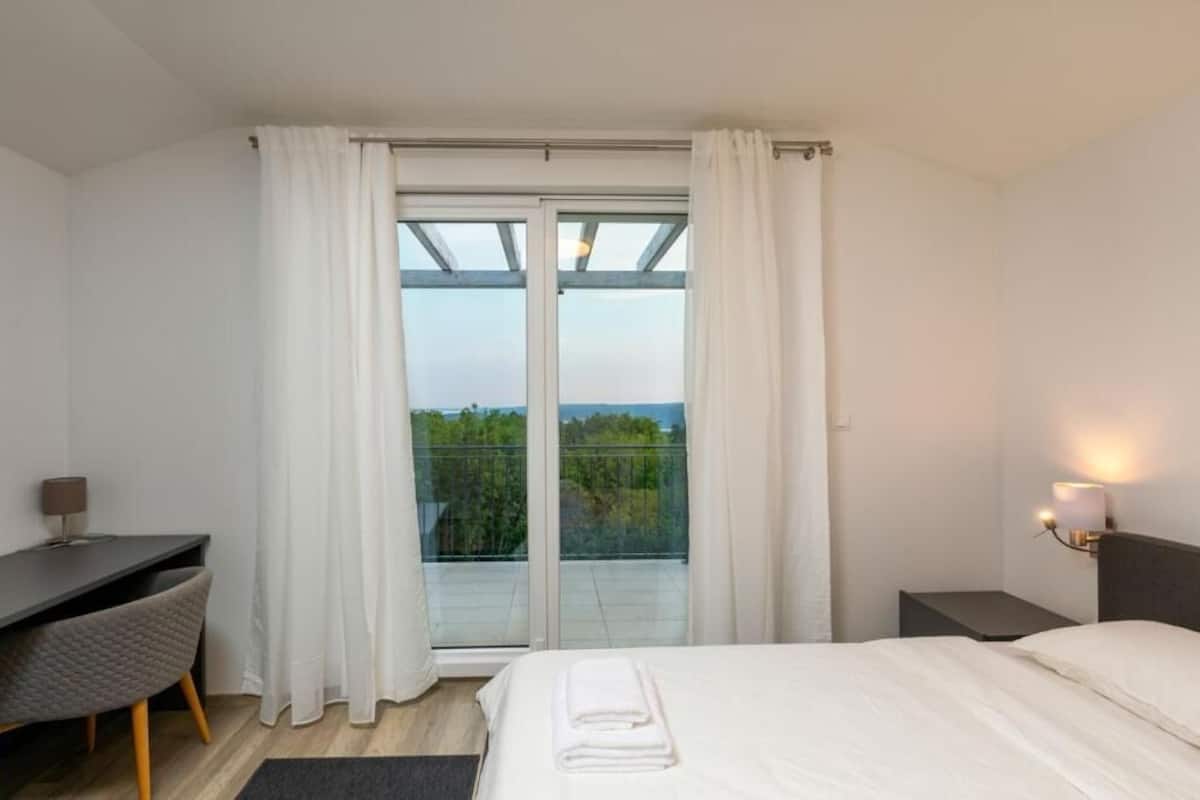 A hotel room with a bed, a desk, a chair, a nightstand, and a view through a glass door to the outdoors.