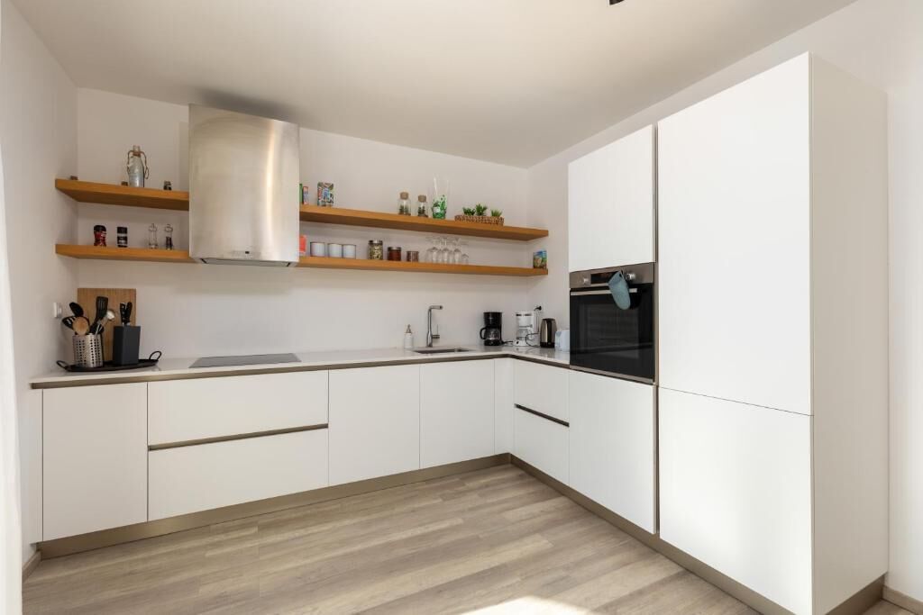 A modern kitchen with white cabinets, wooden shelves, and a stainless steel range hood.