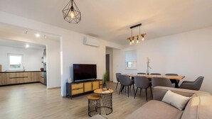 Uma sala de estar moderna com sofá, televisão, área de jantar com cadeiras e cozinha ao fundo.