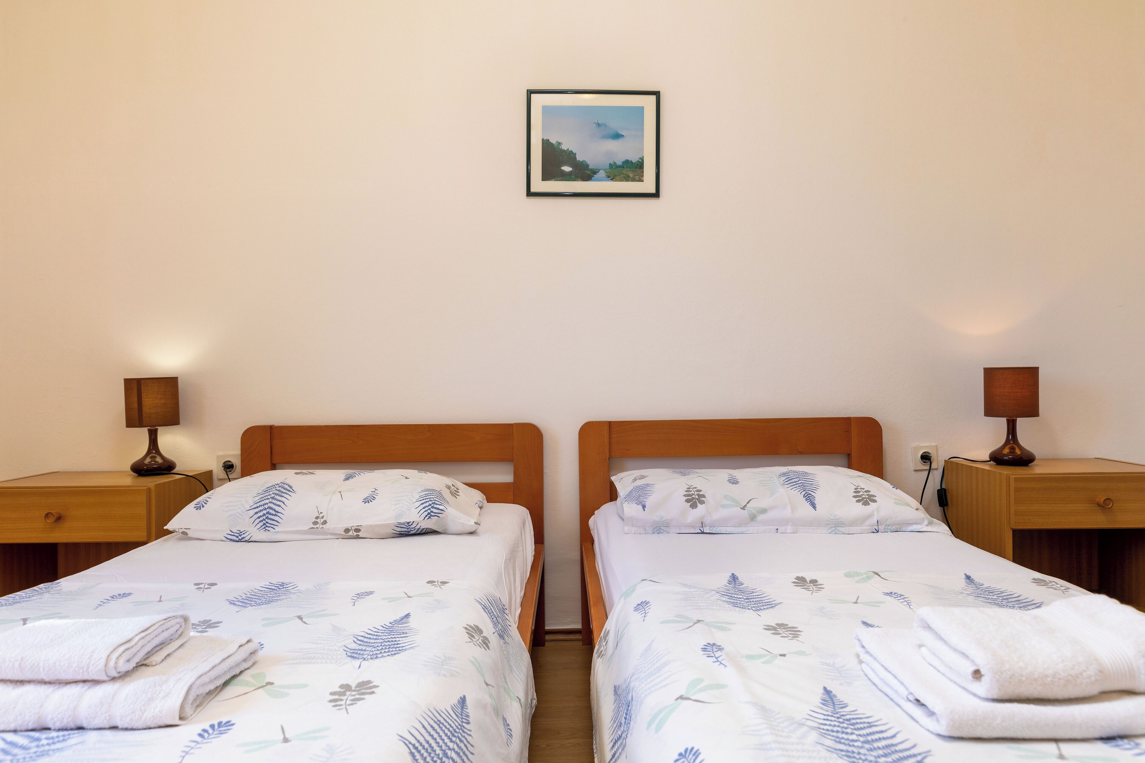 Ein Doppelzimmer mit zwei Einzelbetten, jeweils mit Holz Kopfteil, weißer Bettwäsche mit Blattmuster, Nachttischlampen und einem gerahmten Bild an der Wand.