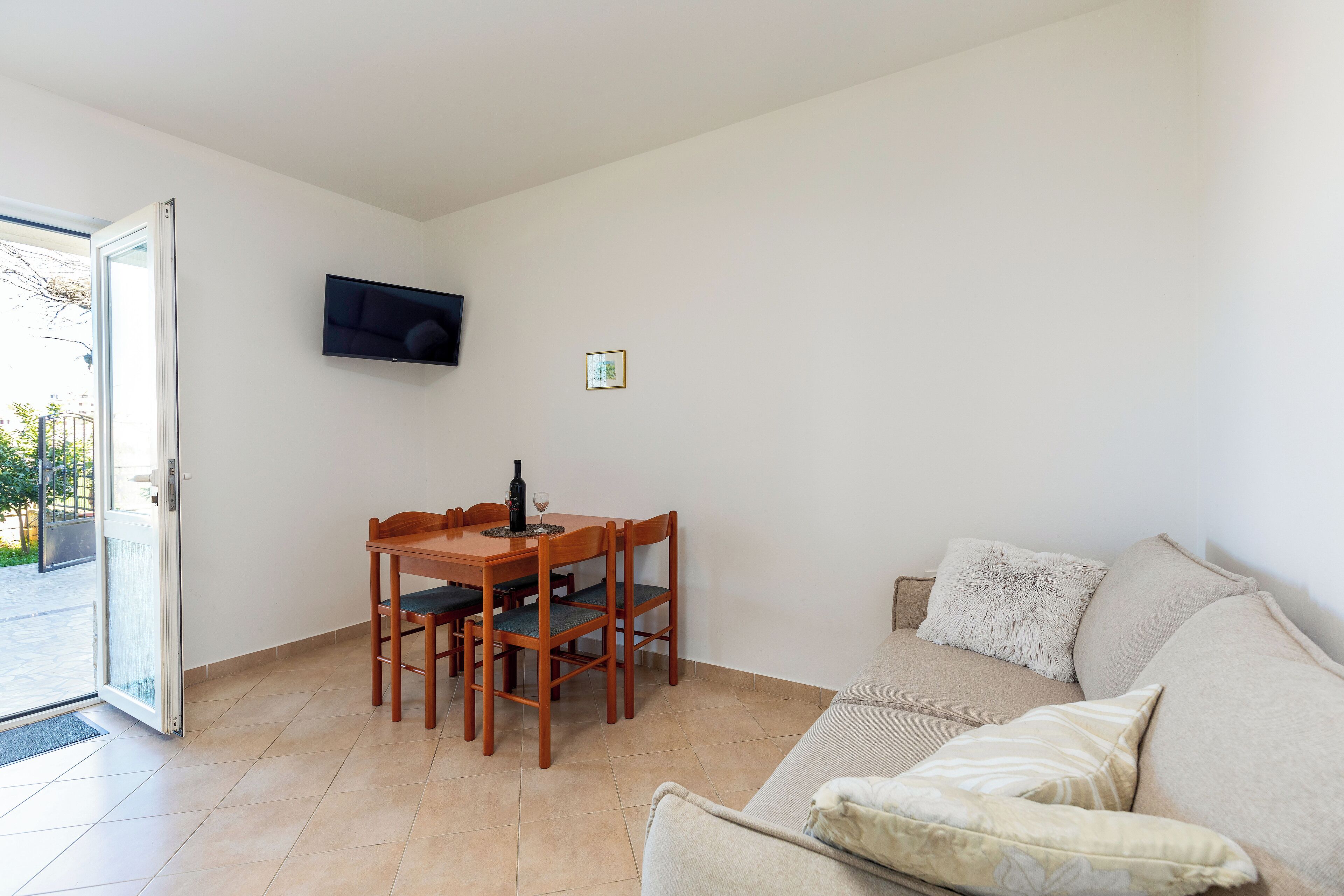 A living room with a sofa, a dining table with chairs, a television mounted on the wall, and a door leading outside.