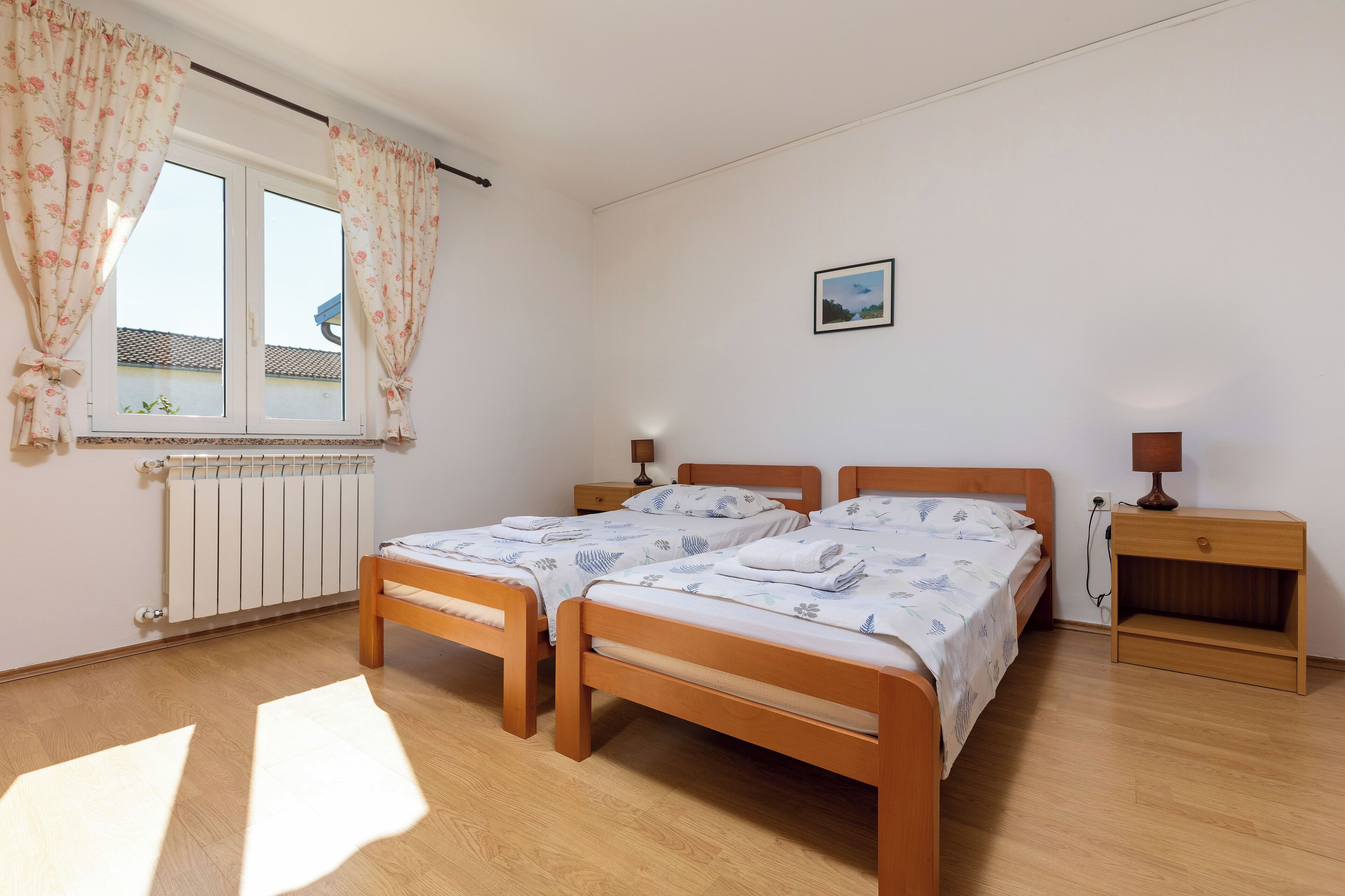 Un dormitorio con dos camas de madera, un radiador, una ventana con cortinas, una mesita de noche con una lámpara y un cuadro pequeño en la pared.