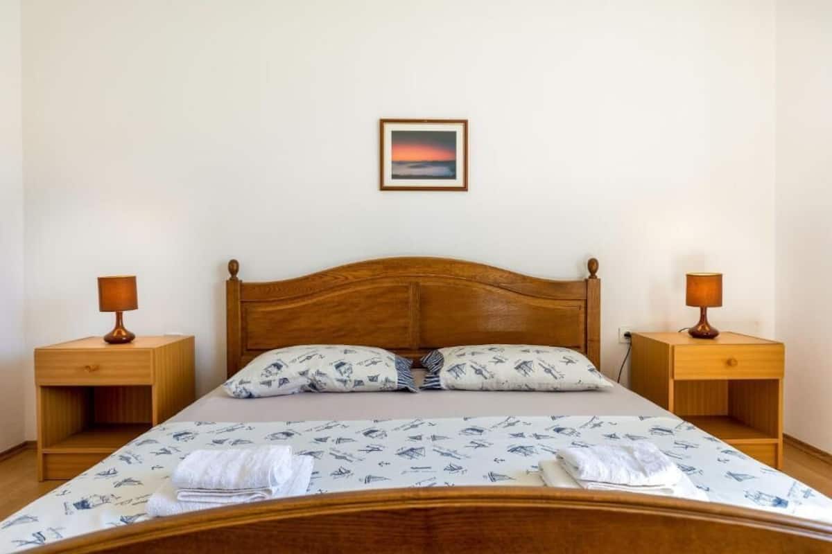 Un lit simple avec une tête de lit en bois, deux tables de chevet avec des lampes, un oreiller à motifs nautiques et un tableau encadré accroché au mur.