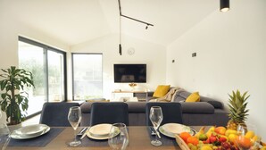 Uma sala de estar moderna com área de jantar, apresentando um sofá, uma TV de tela plana e uma cesta de frutas na mesa.