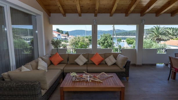 A modern living room with a wooden ceiling, a beige sofa with orange cushions, and a view of a coastal landscape through large windows. - House Kaliope (Zadar)