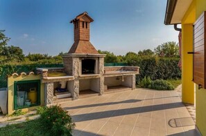 Un'area barbecue esterna con forno a mattoni e canna fumaria, un piccolo spazio chiuso con una porta verde e un edificio giallo sullo sfondo.
