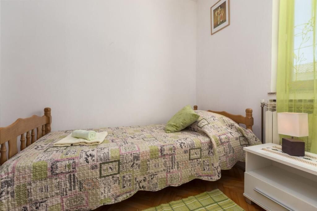 A small bedroom with a single bed, a bedside table with a lamp, and a window with sheer curtains.