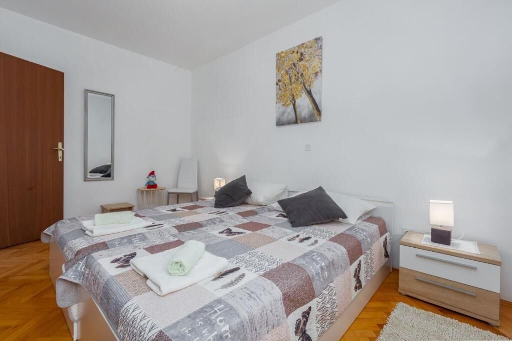 A bedroom with a large bed, a bedside table with a lamp, a chair, a small table with a decorative item, and a painting on the wall.