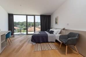 Ein modernes Schlafzimmer mit einem Bett, einem Schreibtisch und einem Stuhl. Ein großes Fenster mit Vorhängen bietet Blick auf Gebäude und Bäume.