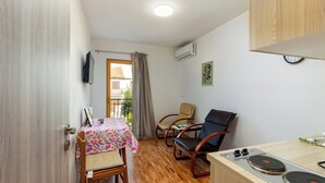 A compact room with a wooden floor, a dining table with chairs, a small kitchenette with a stove, and a window with a curtain.
