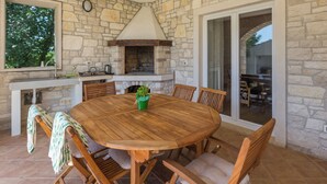 A stone-walled outdoor kitchen with a round wooden table and chairs, a chimney, and a view through a glass door. - Villa Diletta (Kanfanar)