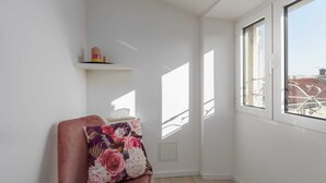 Une pièce d’angle avec un canapé rose, un coussin à motifs floraux, une petite étagère ornée d’objets décoratifs et une grande fenêtre donnant sur des immeubles.