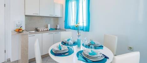 Une cuisine moderne avec des meubles blancs, un évier et un plan de travail sur lequel se trouve un panier de fruits. On y trouve également une table à manger, dressée avec des sets de table bleus et des assiettes blanches.