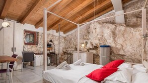 Un dormitorio con pared de piedra, vigas de madera en el techo, una cama con ropa blanca y almohadas rojas, un escritorio con una lámpara y una silla.