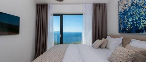 A modern bedroom with a large bed, a view of the sea through a glass door, and a painting on the wall.