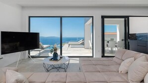 Ein modernes Wohnzimmer mit einem großen Flachbildfernseher, einem beigefarbenen Sofa und einem Couchtisch. Das Zimmer verfügt über große Schiebeglasfenster, die auf einen Balkon mit Blick auf das Meer und die Berge führen.