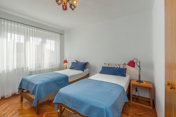 A room with two beds, each with a blue bedspread and pillows, a wooden floor, a window with sheer curtains, and a small table with a lamp.