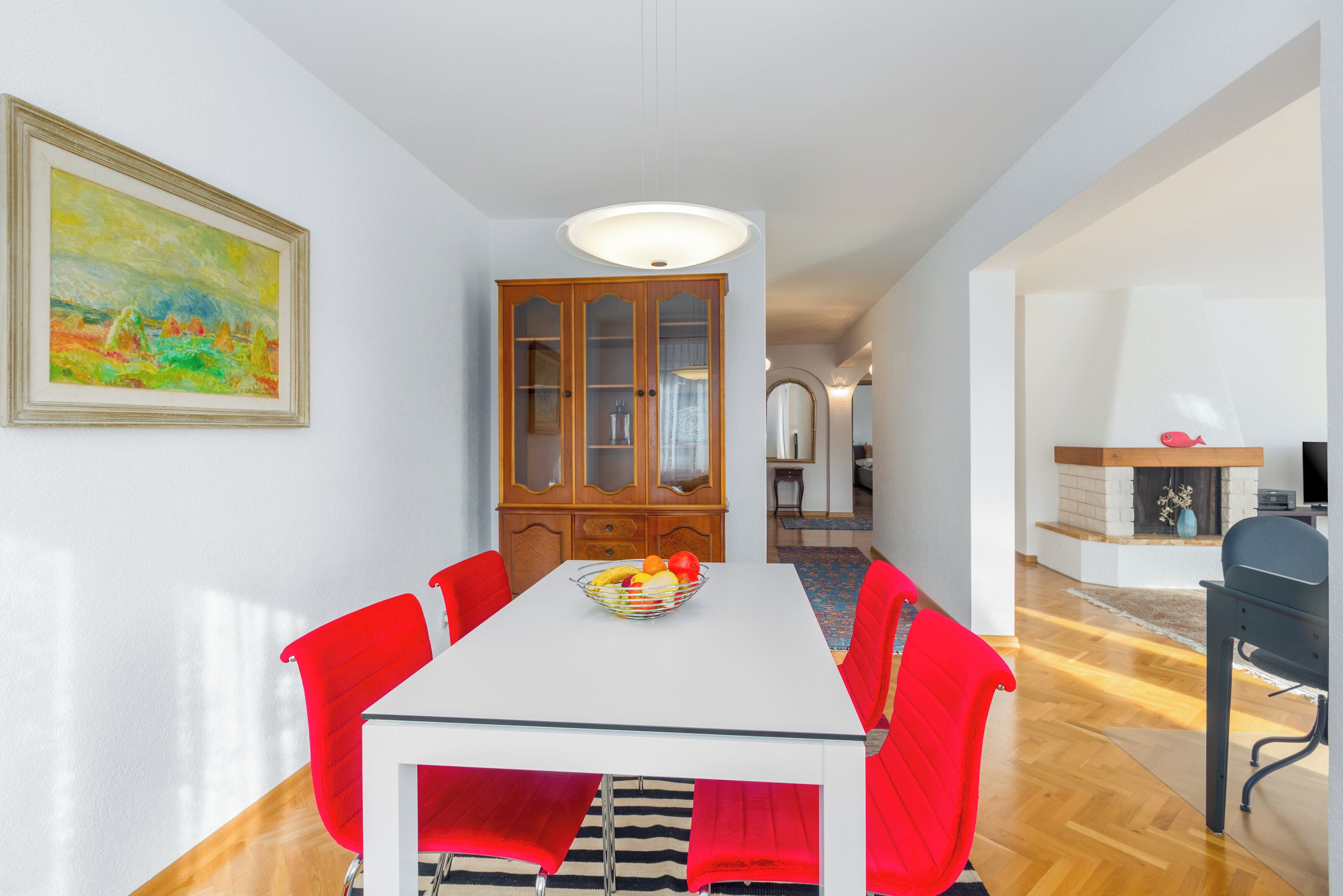 Uma sala de jantar com uma mesa branca, cadeiras vermelhas, um armário de madeira e um quadro na parede.
