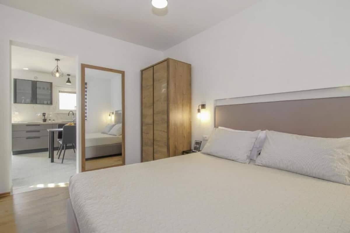 A bedroom with a bed, a wardrobe, a bedside table, and a nightstand. There is a door leading to a bathroom and a kitchenette.