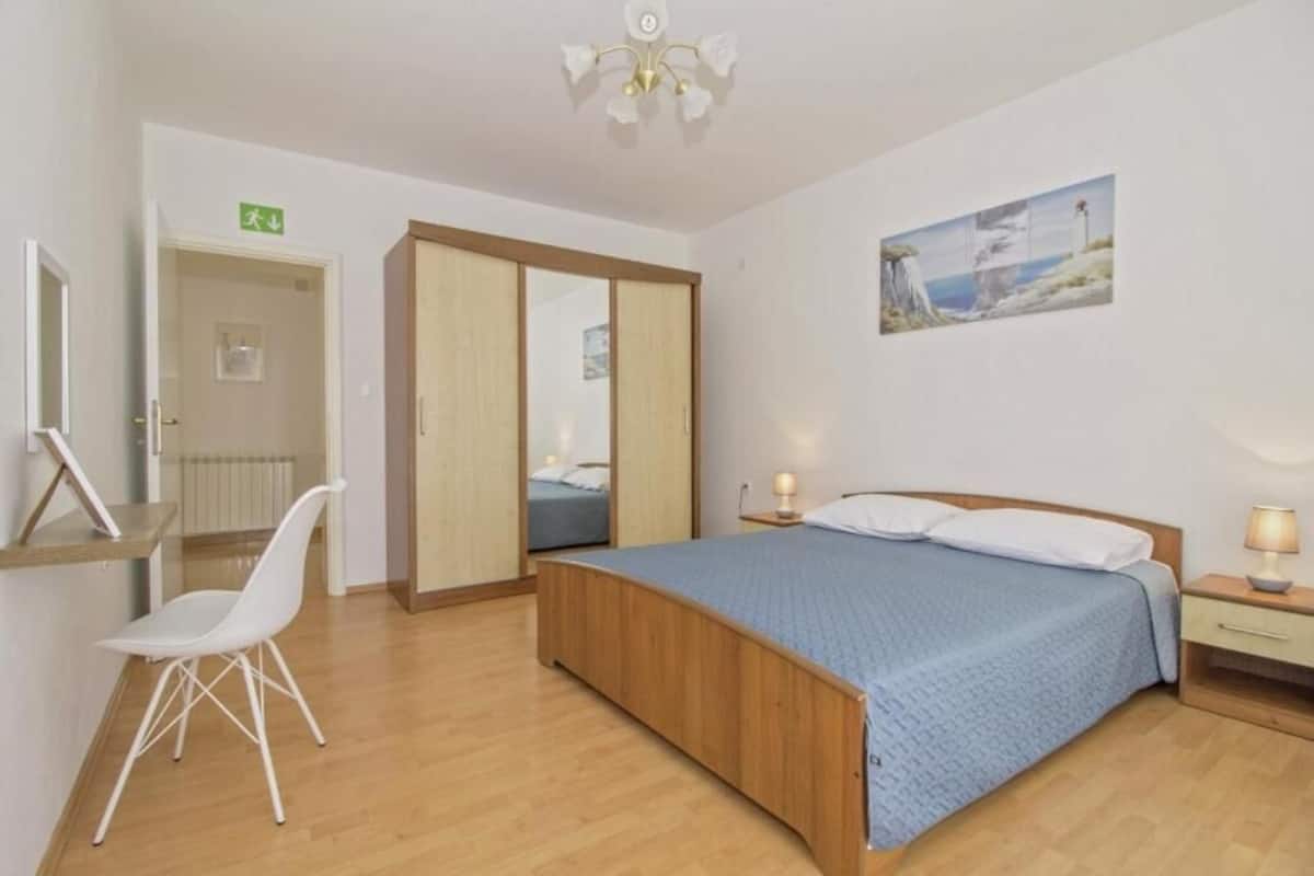 A bedroom with a wooden bed, a white chair, a bedside table with a lamp, and a painting on the wall.