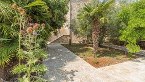 A stone pathway leading to a stone building with a chimney, surrounded by lush greenery and palm trees.