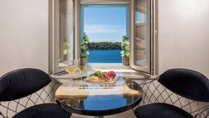 A balcony with a table set for two, featuring a fruit platter and a glass of wine, overlooking a body of water and greenery. - Natasha's Beachfront (Rovinj)