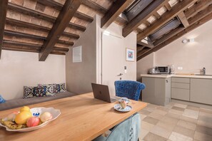 A dining area with a wooden table, a laptop, and a bowl of fruit. There is a blue chair and a sofa with patterned cushions. A kitchen is visible in the background. - Natasha's Beachfront (Rovinj)