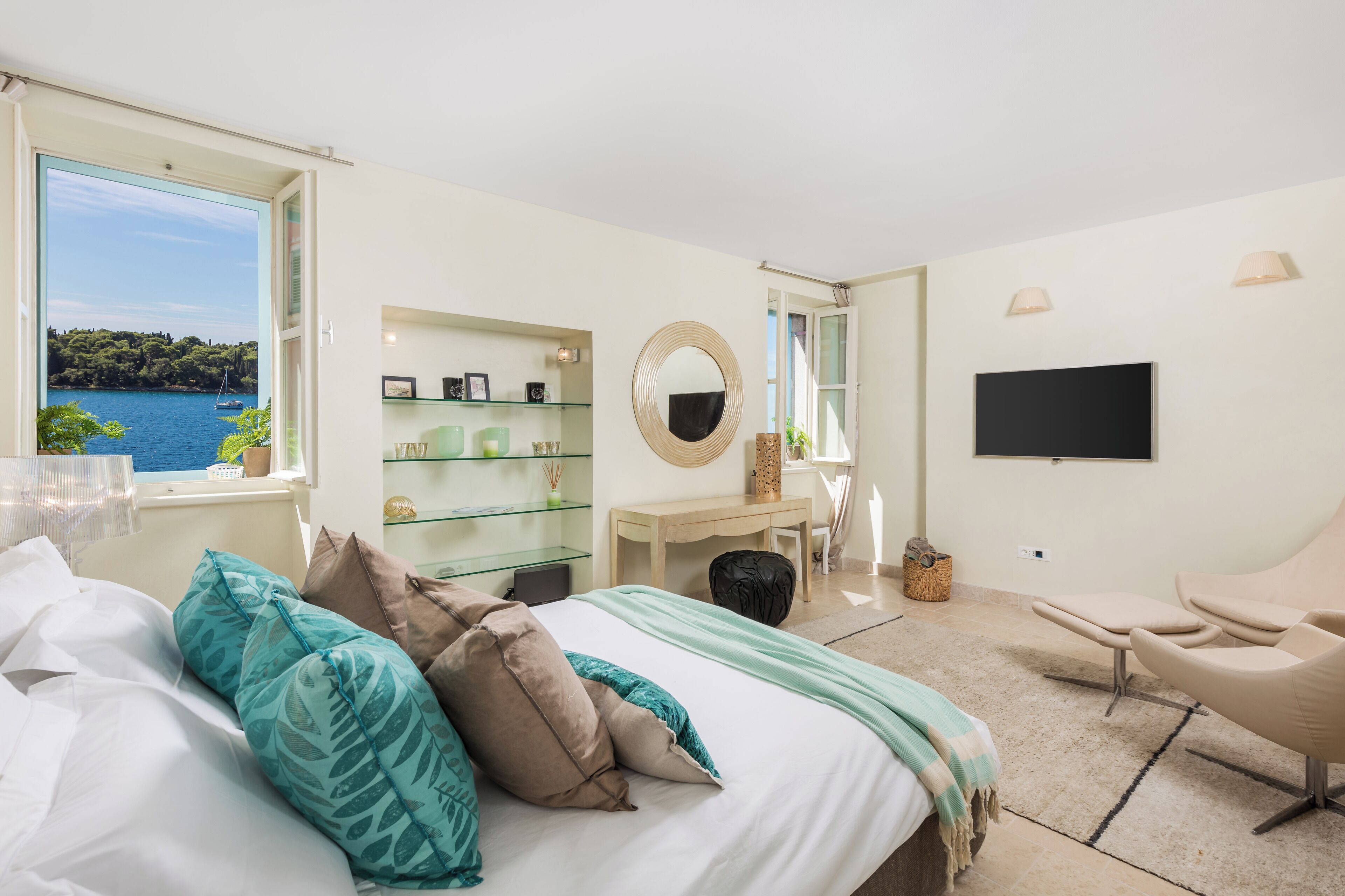 A bedroom with a bed, a chair, a desk, a TV, and a view of the sea.