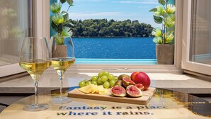 A table with two glasses of white wine, a wooden board with grapes, figs, and cheese, and a view of a lake through a window. - Natasha's Beachfront (Rovinj)
