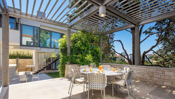 A covered outdoor dining area with a table and chairs, a pergola, and a view of a modern building and trees. - RM Grande Villa (Rovinj)