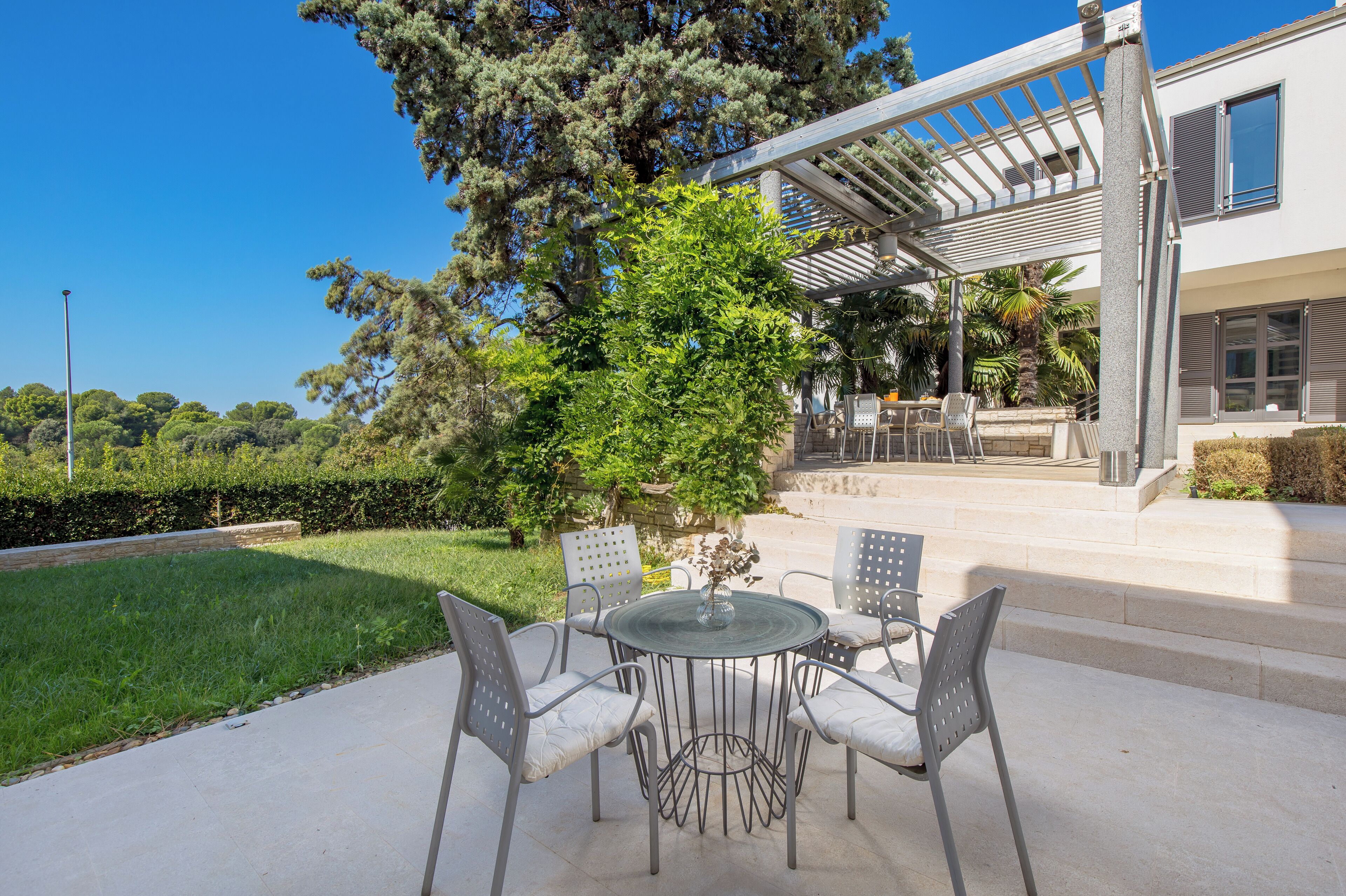 Une terrasse avec une table ronde et des chaises, une pergola et une vue sur une maison avec un jardin.