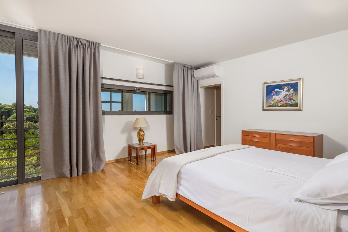 Ein Schlafzimmer mit einem großen Bett, einer Holzkommode, einem Nachttisch mit Lampe, einem Fenster mit Vorhängen und einem Bild an der Wand.