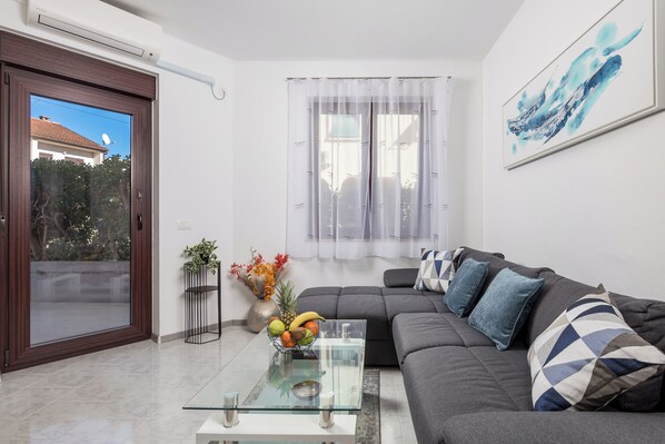 Una sala de estar moderna con una mesa de centro de vidrio, un sofá gris con cojines estampados, una pared blanca con un cuadro abstracto enmarcado y un ventanal corredero que da a un espacio exterior.