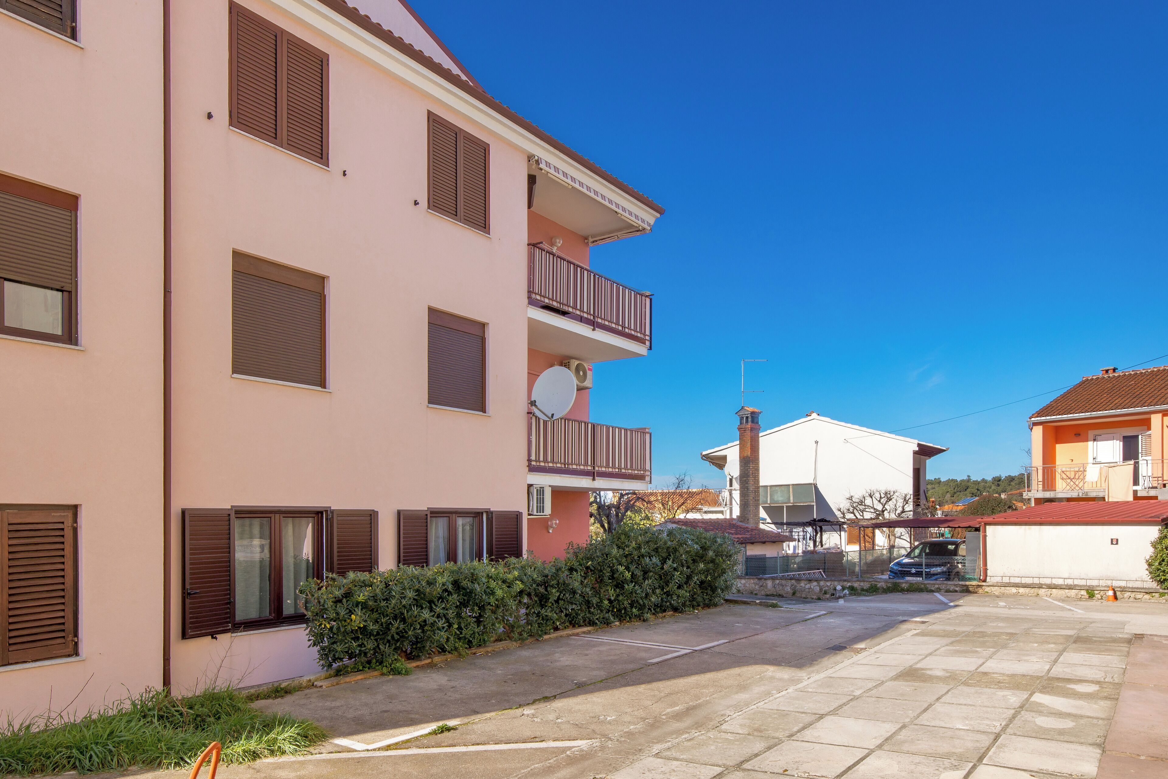 A multi-story residential building with balconies, a parking area, and a clear blue sky.
