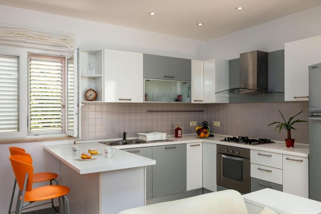 Une cuisine moderne avec des meubles blancs, une cuisinière à gaz et un évier. On y trouve deux chaises orange et une table blanche.