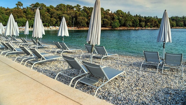 Una zona sulla spiaggia con file di lettini e ombrelloni, una spiaggia di ciottoli e un mare calmo con una costa alberata sullo sfondo.