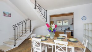 Un comedor con una mesa de madera dispuesta para cuatro, sillas blancas y un florero con flores. Una escalera con barandilla negra conduce a un piso superior.