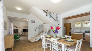 Amplia sala de estar con una mesa de comedor de madera, sillas blancas, una escalera y una zona de cocina.